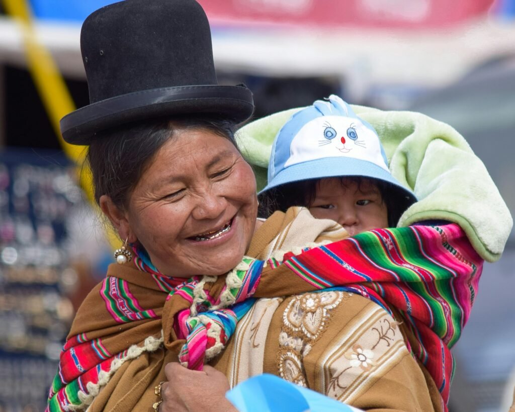 pexels-photo-17028097-17028097 A joyful Bolivian woman in traditional clothing carrying a child in a vibrant shawl, outdoors.